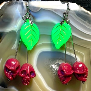 May include: A pair of unique earrings featuring green leaf-shaped charms with silver hooks. Below each leaf hangs a cluster of three red skull-shaped charms. The earrings are designed with a dark, gothic aesthetic.