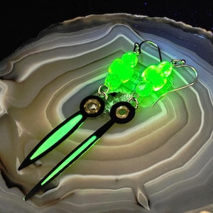 May include: A pair of unique earrings featuring a black and neon green design. The earrings have a circular black top with a gold center and a long, pointed neon green bottom. The earrings also have a cluster of bright green beads at the top.