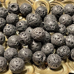May include: A collection of dark gray, porous lava rock beads. The spherical beads have a rough, textured surface with numerous small holes. They are displayed on a gold-colored, ornate dish, creating a contrast in textures and colors.