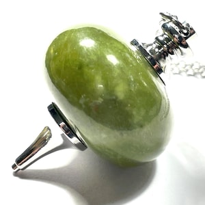 May include: A green jade pendant with a silver-colored metal clasp and a matching silver chain. The jade is a smooth, oval shape. The pendant's clasp is intricately designed. The pendant is displayed against a white background.