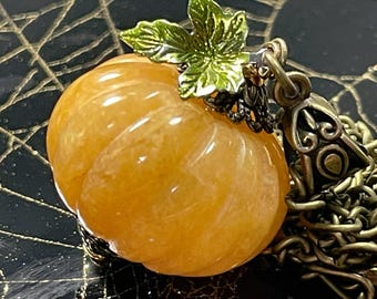 May include: A pumpkin-shaped pendant necklace. The orange gemstone pumpkin has carved ridges and is topped with a green leaf and a decorative metal clasp. The necklace chain is a bronze color.