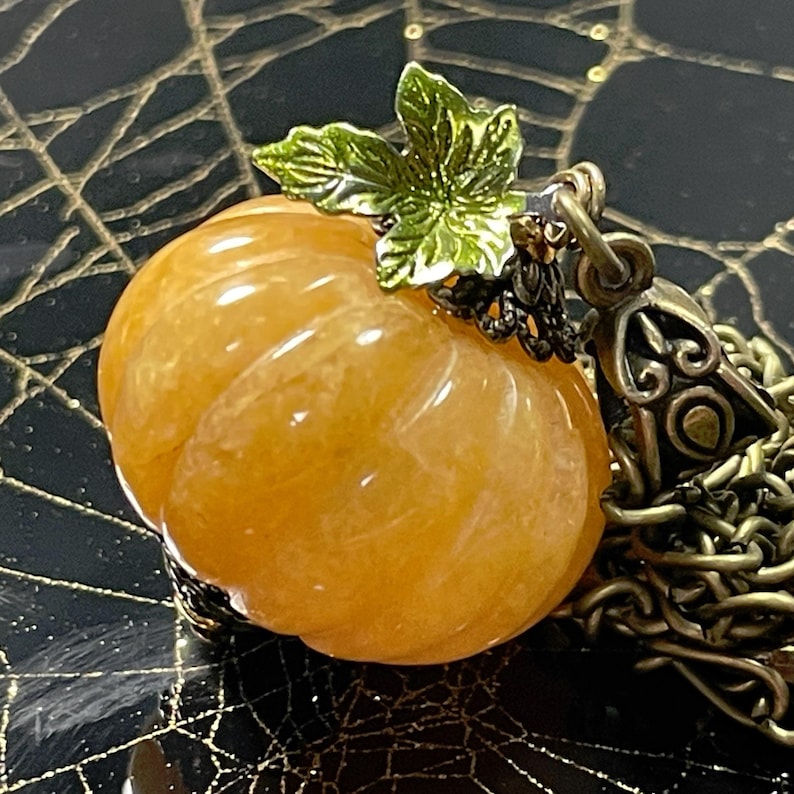 May include: A pumpkin-shaped pendant necklace. The orange gemstone pumpkin has carved ridges and is topped with a green leaf and a decorative metal clasp. The necklace chain is a bronze color.