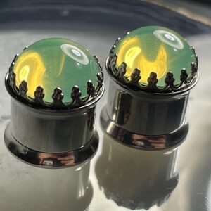 May include: A pair of green glass ear gauges with silver metal settings. The gauges have a decorative crown design around the glass.