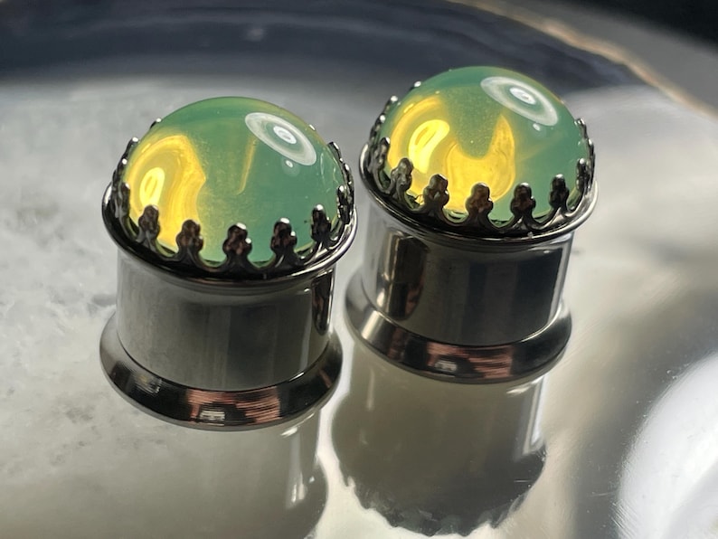 May include: A pair of green glass ear gauges with silver metal settings. The gauges have a decorative crown design around the glass.
