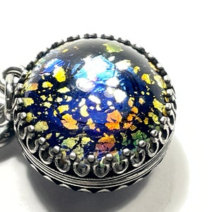 May include: A round pendant with a dark blue, glittery surface, accented with gold and other colorful flecks. The pendant is encased in a silver-colored, ornate metal setting. The design evokes a celestial or cosmic theme.