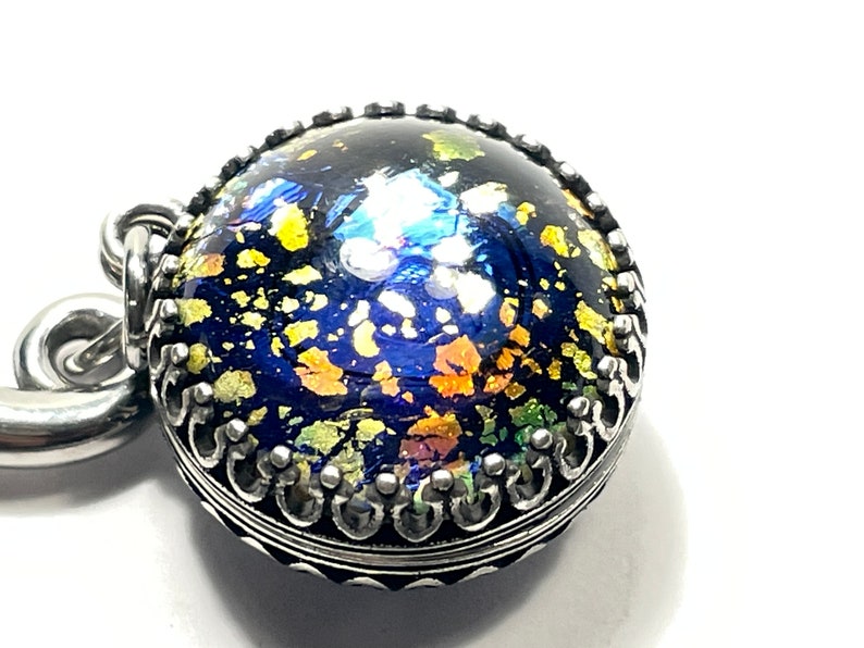 May include: A round pendant with a dark blue, glittery surface, accented with gold and other colorful flecks. The pendant is encased in a silver-colored, ornate metal setting. The design evokes a celestial or cosmic theme.