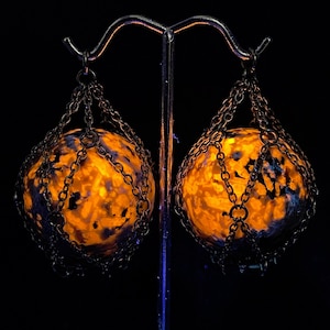 Puede incluir: Un par de pendientes únicos con piedras esféricas brillantes en tonos naranja y azul, encerradas en una jaula de cadena metálica. Los pendientes cuelgan de un soporte de metal plateado sobre un fondo negro.
