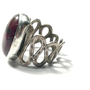 May include: A silver ring with a large, oval-shaped, red gemstone set in a bezel setting. The band of the ring is intricately designed with a series of connected loops.