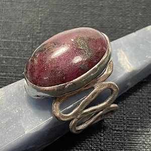May include: A ring featuring a large, oval-shaped, deep maroon cabochon stone with dark inclusions, set in a silver-toned metal bezel. The band is constructed from a continuous, wavy, textured silver-toned wire that loops to form the ring structure.