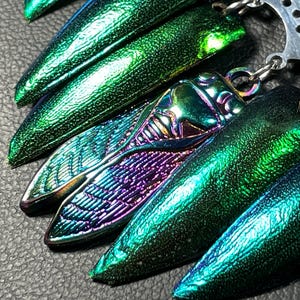 May include: Close-up of several iridescent cicada wing pendants in shades of green, teal, and purple. The pendants are attached to a silver chain. The wings have a textured, metallic finish.