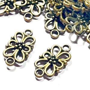 May include: Close-up of several antique brass-colored filigree flower charms. Each charm features a floral design with a central cluster of small beads and looped petals. The charms have small loops for attaching to other jewelry components.