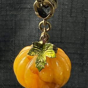 May include: A gold-toned necklace pendant featuring a small, orange pumpkin with a green leaf. The pumpkin has a ribbed texture and is suspended from a gold chain. The pendant is likely a seasonal or autumn-themed accessory.