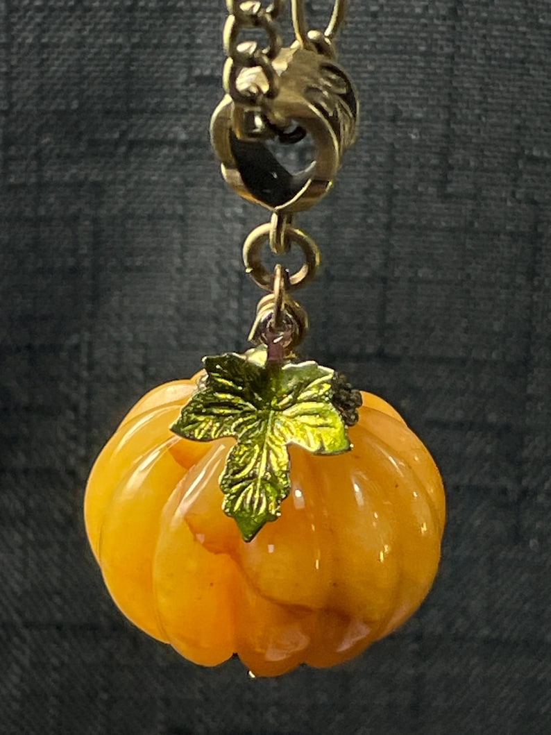 May include: A gold-toned necklace pendant featuring a small, orange pumpkin with a green leaf. The pumpkin has a ribbed texture and is suspended from a gold chain. The pendant is likely a seasonal or autumn-themed accessory.