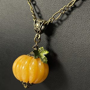 May include: A gold-toned necklace featuring a pendant shaped like a small, orange pumpkin. The pumpkin has carved vertical lines and is topped with a green leaf and a decorative metal cap. The chain is a linked design.