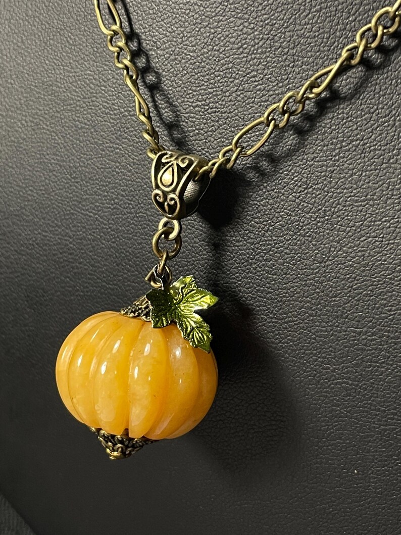 May include: A gold-toned necklace featuring a pendant shaped like a small, orange pumpkin. The pumpkin has carved vertical lines and is topped with a green leaf and a decorative metal cap. The chain is a linked design.