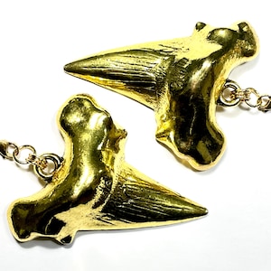 May include: Two gold-coloured shark tooth pendants with attached gold-tone chains and clasps. The pendants are highly detailed, with a textured surface and a polished finish. The background is white.