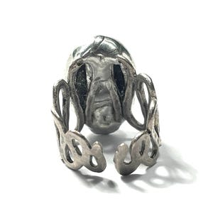 May include: A silver ring with a detailed skull design. The ring is open and adjustable.