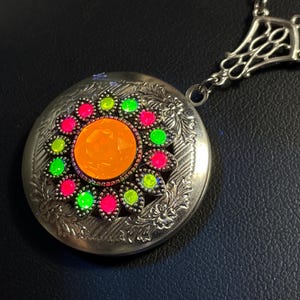 May include: Silver-toned locket necklace with a floral design. The locket features a central orange gem surrounded by pink and green gems. The necklace has a decorative chain.