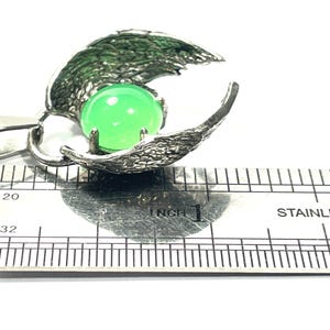 K&ouml;nnte beinhalten: Silberne Halskette mit Anh&auml;nger, der einen gr&uuml;nen Edelstein in einem detaillierten Fl&uuml;geldesign tr&auml;gt. Der Anh&auml;nger hat einen Durchmesser von etwa 2,5 cm.