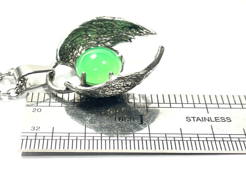 K&ouml;nnte beinhalten: Silberne Halskette mit Anh&auml;nger, der einen gr&uuml;nen Edelstein in einem detaillierten Fl&uuml;geldesign tr&auml;gt. Der Anh&auml;nger hat einen Durchmesser von etwa 2,5 cm.
