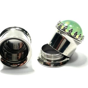 May include: A pair of stainless steel ear gauges with a green glass insert. The insert is set in a silver crown-like setting. One gauge is fully assembled, while the other is disassembled, showing the individual pieces.