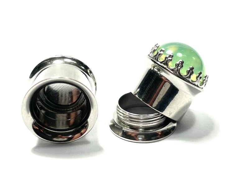 May include: A pair of stainless steel ear gauges with a green glass insert. The insert is set in a silver crown-like setting. One gauge is fully assembled, while the other is disassembled, showing the individual pieces.