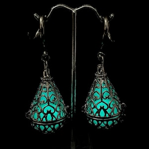May include: A pair of dark metal drop earrings featuring an intricate filigree cage design in a teardrop shape. Inside each metal cage is a glowing, luminous turquoise orb that emits a bright light against the dark, detailed metal framework.