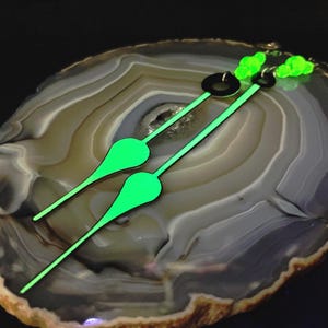 May include: Two watch hands with a neon green glow. The hands have a black outline and a teardrop-shaped tip. The hands are set against a background of a polished, multi-toned stone.