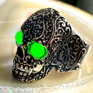 May include: A silver skull ring with intricate details and green glowing eyes. The ring is set against a white background.