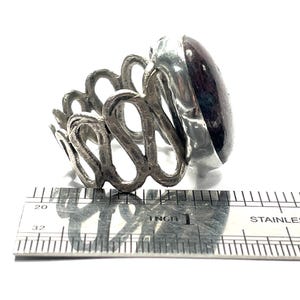 May include: A silver ring with an intricate design and a large, oval-shaped, dark red gemstone. The ring is measured against a ruler, showing it is approximately 1 inch in diameter.