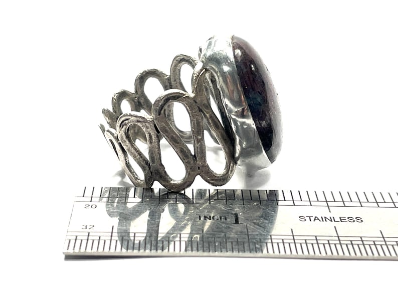 May include: A silver ring with an intricate design and a large, oval-shaped, dark red gemstone. The ring is measured against a ruler, showing it is approximately 1 inch in diameter.
