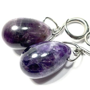May include: Two polished amethyst gemstones with silver-colored metal loops. The gemstones are oval-shaped and display a range of purple hues. The background is white, highlighting the gemstones' color and texture. The gemstones are connected by a silver chain.