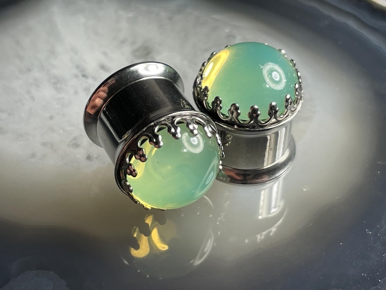 May include: A pair of silver ear gauges with a green stone inlay and a decorative crown design.