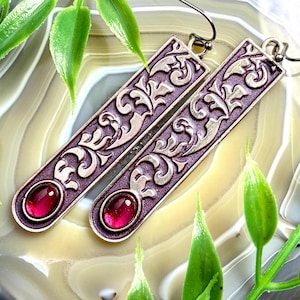 May include: A pair of silver-toned earrings with a rectangular design and a deep purple background. The earrings feature an ornate, swirling pattern and a small, oval-shaped, red gemstone at the bottom. The earrings are displayed on a light-coloured, patterned surface with green leaves.