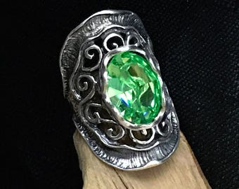 May include: A silver ring featuring a large, oval-cut, vibrant lime green gemstone set in the center. The metal band displays an intricate, dark, oxidized filigree pattern with a textured, scalloped edge surrounding the central stone.