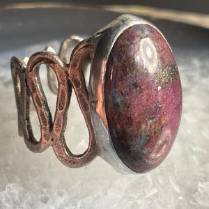 May include: A silver ring with a pink and grey oval gemstone set in a silver bezel. The band is made of copper and has a textured, organic design.
