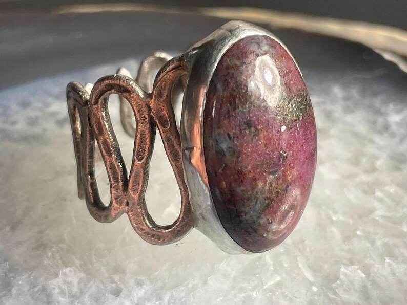 May include: A silver ring with a pink and grey oval gemstone set in a silver bezel. The band is made of copper and has a textured, organic design.