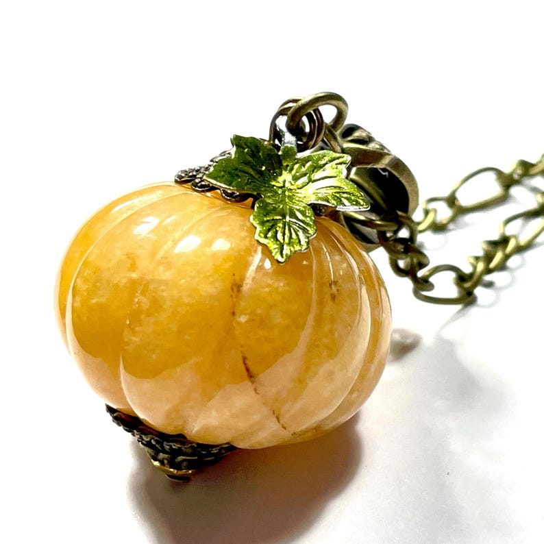 May include: A pumpkin-shaped pendant necklace. The pendant is a light orange color with a glossy finish, and is accented with a green leaf and antique bronze-colored metal. The chain is also antique bronze-colored.