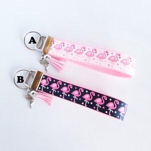 May include: Two pink flamingo keychains with silver key rings and pink tassels. The keychain on the top has a pink ribbon with white polka dots and pink flamingos. The keychain on the bottom has a black ribbon with white polka dots and pink flamingos. The keychains are labeled "A" and "B".