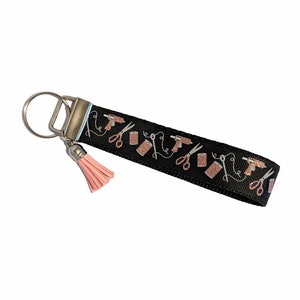 May include: Black key chain with a pink tassel. The key chain has a white and rose gold design of scissors, glue guns, and thread.