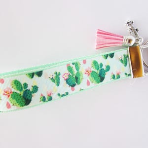 May include: A light blue wristlet keychain with a repeating pattern of green cacti and pink flowers. The keychain has a silver-colored metal clasp, a pink tassel, and a metal key ring.
