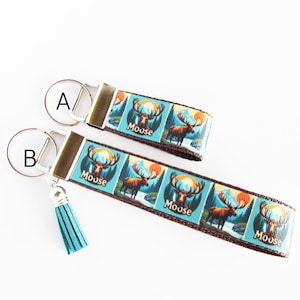 May include: Two keychains with a blue and brown ribbon featuring a moose design and the word "Moose". One keychain has a silver ring and the other has a blue tassel. The background is white.