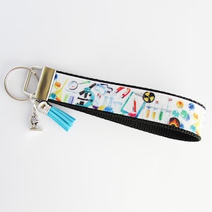 May include: A white and black key chain with a blue tassel. The white part of the key chain has a colorful print of science-related items, including beakers, test tubes, and a microscope.