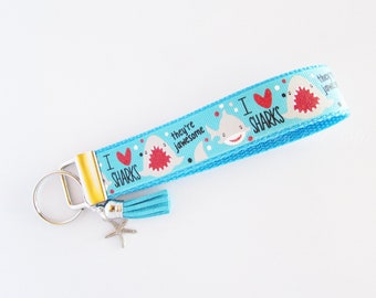 May include: A blue and white key chain with a shark design. The key chain has a tassel and a silver key ring. The text on the key chain reads "I love sharks, they're jawsome".
