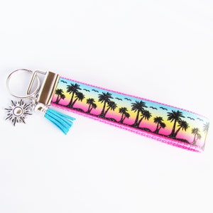 May include: A pink key chain with a silver key ring and a blue tassel. The key chain features a colorful sunset design with palm trees and a silver sun charm.