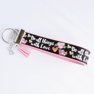 May include: A black and pink keychain wristlet with the text "Do all things with Love" and floral designs. The wristlet has a pink tassel and a silver heart charm. The keychain is on a silver metal ring.