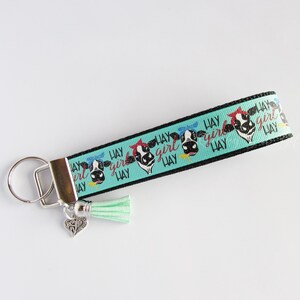 May include: A turquoise and black key chain with a cow print design and the text "Hay Girl Hay". The key chain has a silver key ring and a small silver charm with a cow design. A turquoise tassel hangs from the bottom of the key chain.
