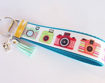 May include: A teal blue key chain with a silver key ring and a light blue tassel. The key chain has a white ribbon with four colorful camera designs printed on it.
