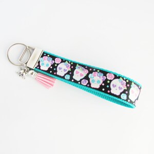 May include: A turquoise and black key chain with a pink tassel and a silver skull charm. The key chain features a repeating pattern of white sugar skulls with pink, purple, and green flowers.