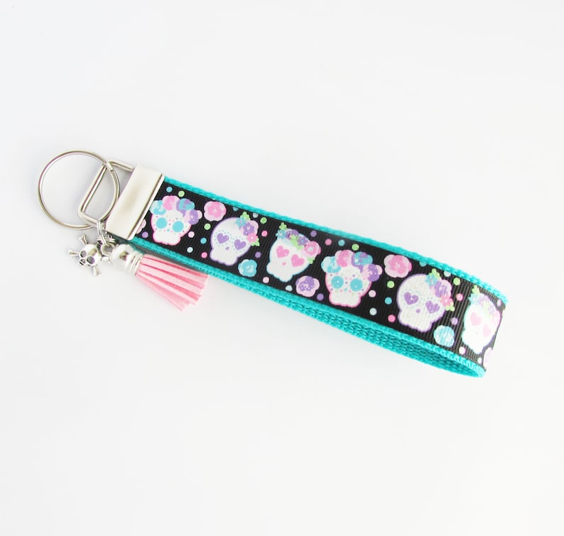 May include: A turquoise and black key chain with a pink tassel and a silver skull charm. The key chain features a repeating pattern of white sugar skulls with pink, purple, and green flowers.